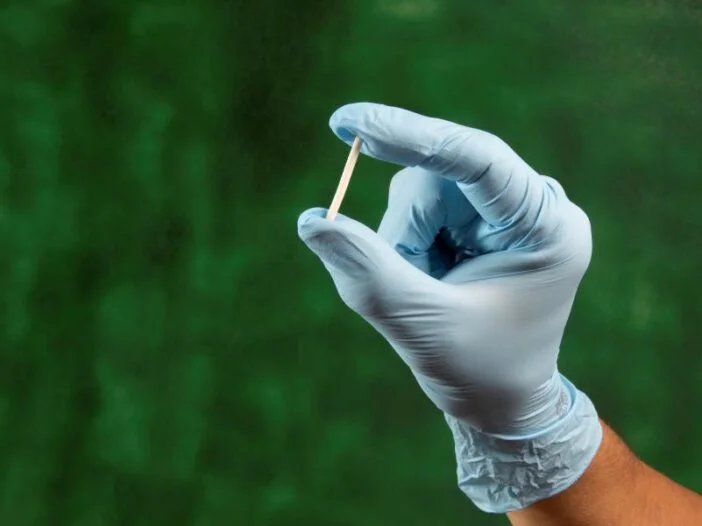 ão com luva cirúrgica segurando um implante hormonal sobre fundo verde