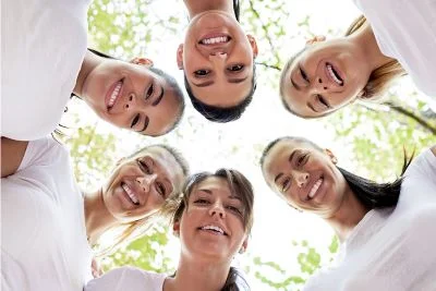 Rosto de seis mulheres de diferentes idades formando um círculo vistas de baixo para cima com natureza ao fundo