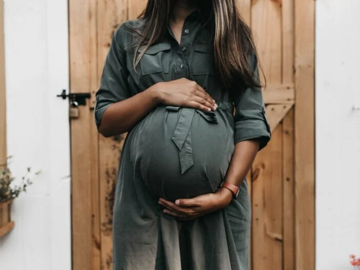 O Mistério da Bolsa das Águas e Seu Rompimento - imagem de mulher grávida com as mãos acima e abaixo da barriga, em vestido verde com porta de madeira atrás